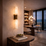 The Eryon LED Small Alabaster Wall Light in brushed brass and soft white glows warmly above a console with books and a bowl, while contemporary dining furniture frames a city view at dusk in the background.