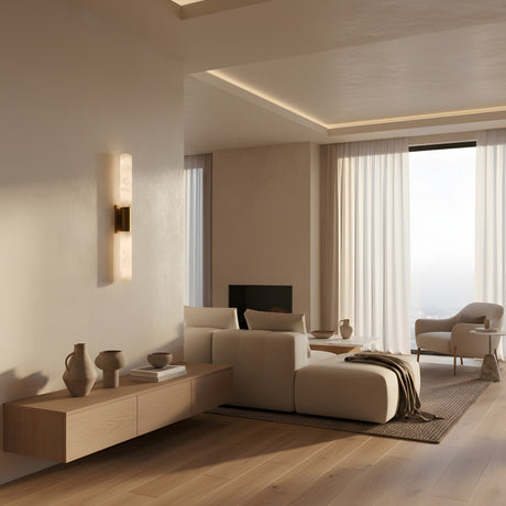 A modern living room in neutral tones with a white sofa, minimalist wood furniture, ceramic vases, and sheer-curtained large windows softly lit by the Eryon LED Small Alabaster Wall Light – Brushed Brass & Soft White.