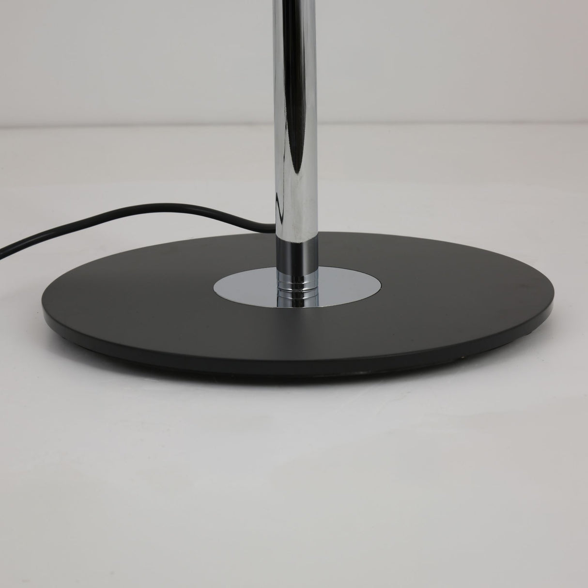 Close-up of the Niori Stealth floor lamp's round matt black metal base with polished chrome stem rising from a central disc, on a plain background.