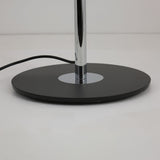 Close-up of the Niori Stealth floor lamp's round matt black metal base with polished chrome stem rising from a central disc, on a plain background.