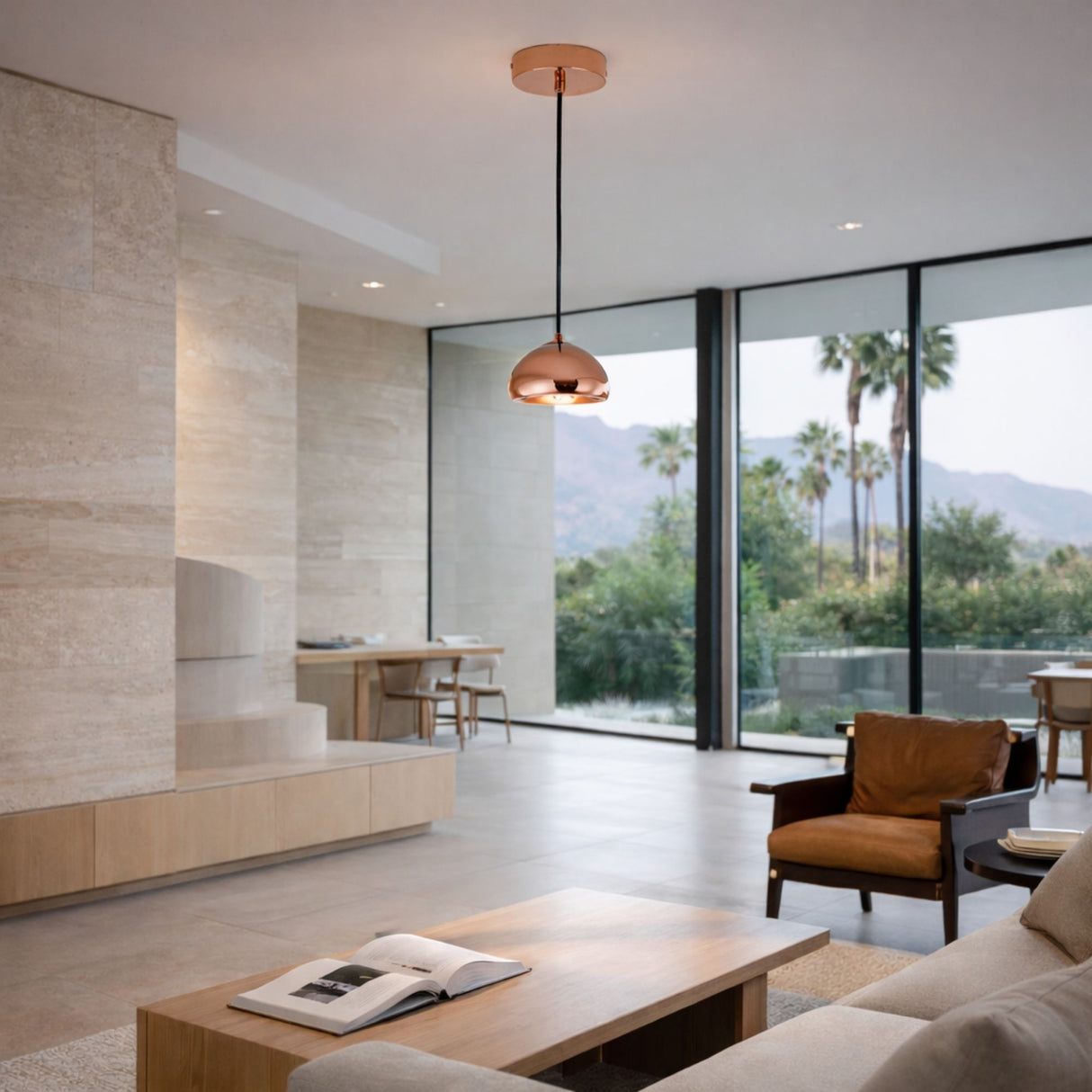 Modern living room with large glass windows, minimal decor, light stone walls, neutral furniture, a wooden coffee table with an open book, and the Grand Ball Glass Pendant 25cm - Copper illuminating views of palm trees and mountains outside.