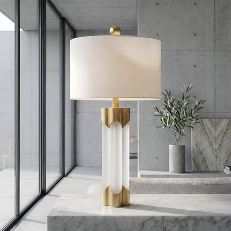 The Lunivar 1 Light Faceted Column Alabaster Table Lamp in brass and soft white sits on a marble surface in a minimalist room with large windows, concrete walls, and a potted plant.