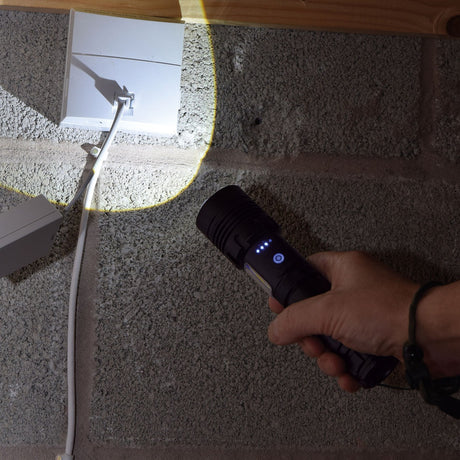 Hand holding the Mercury AVSL rechargeable LED torch with lit side lamp and blue power button, illuminating a socket and cable on a concrete wall.