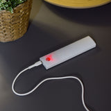 LYYT motion sensing LED light in white plastic casing charging via USB-C cable with red indicator lit, on a dark surface beside a woven planter.