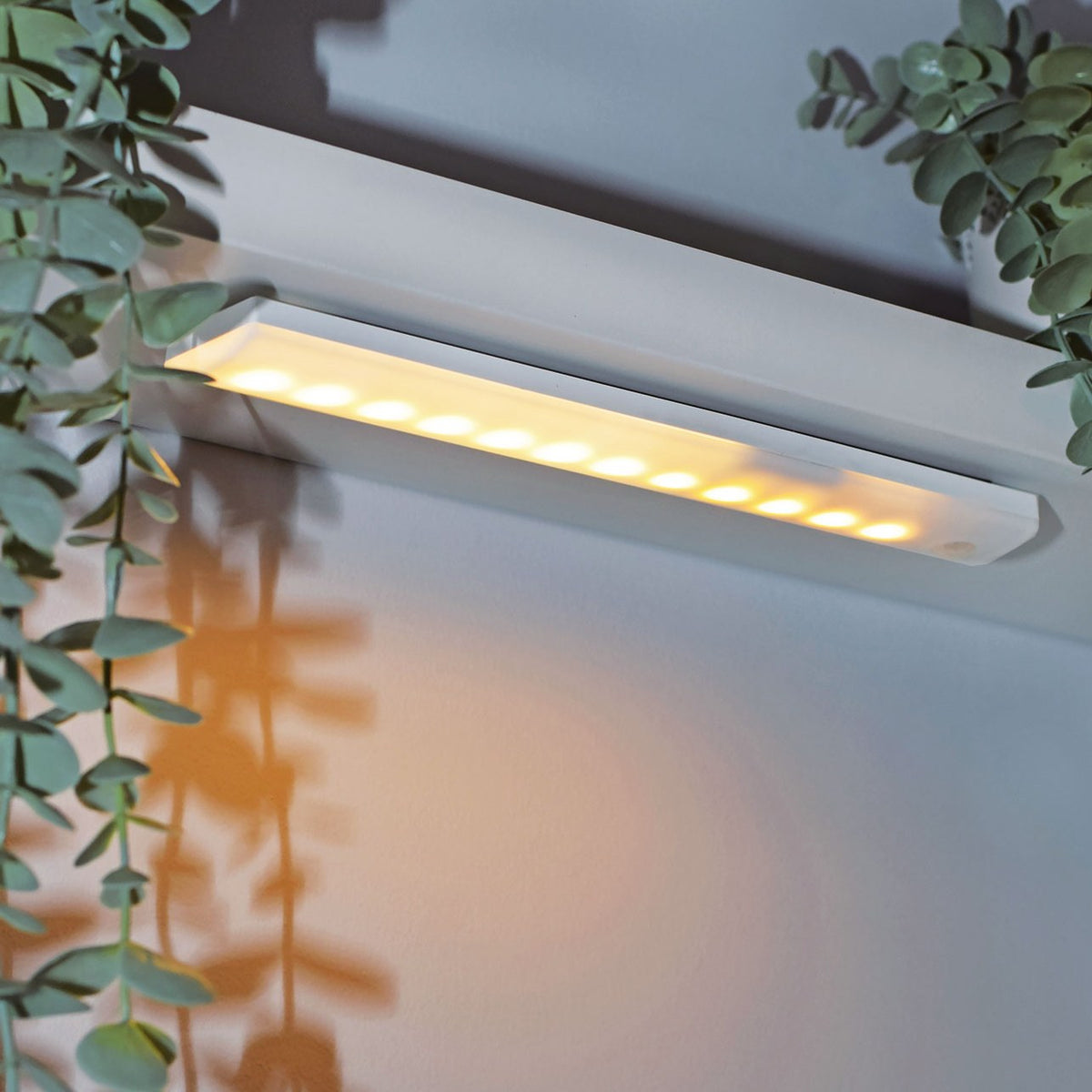 LYYT 36 LED motion sensing light bar by AVSL, mounted under a shelf with warm illumination on a white wall beside trailing eucalyptus foliage.