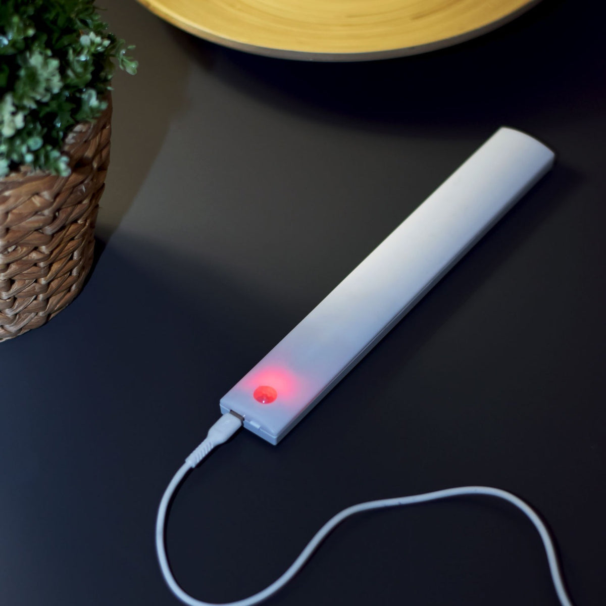 LYYT slim white LED motion sensing light bar charging via USB-C with red indicator light, on a dark surface beside a wicker plant pot.