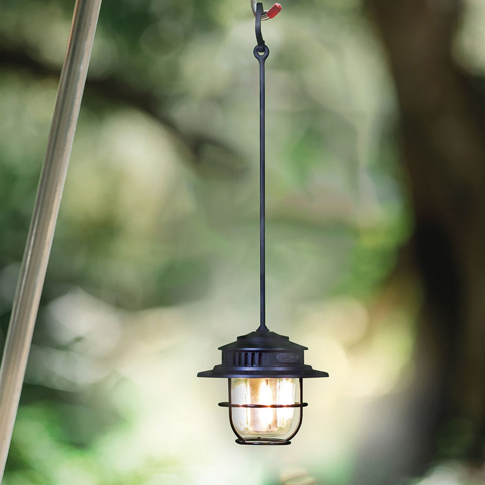AVSL LYYT Rechargeable LED Outdoor Lantern in matt black, hanging by its hook and cord from a tripod pole against a blurred green foliage background.