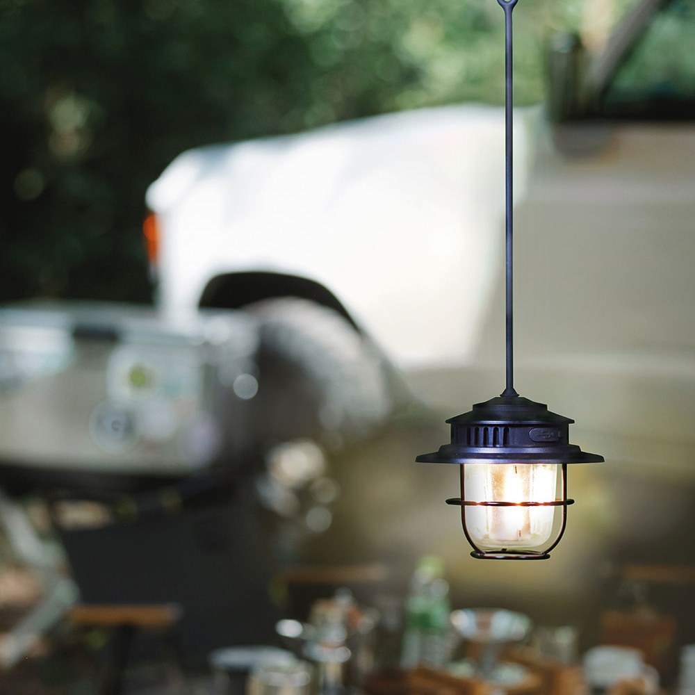 LYYT Rechargeable LED Outdoor Lantern in matt black hanging by its cord at a campsite, with wire cage around the clear lens glowing warm white.