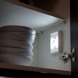 LYYT AVSL LED battery switch light in white ABS with centre rocker switch illuminated inside a kitchen cupboard beside stacked white bowls.