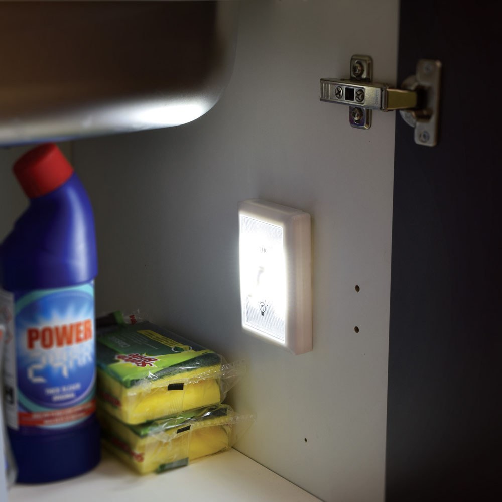 LYYT LED Battery Switch Light by AVSL, white rectangular panel illuminated inside a kitchen cupboard beside cleaning bottle and sponges, mounted to rear wall.