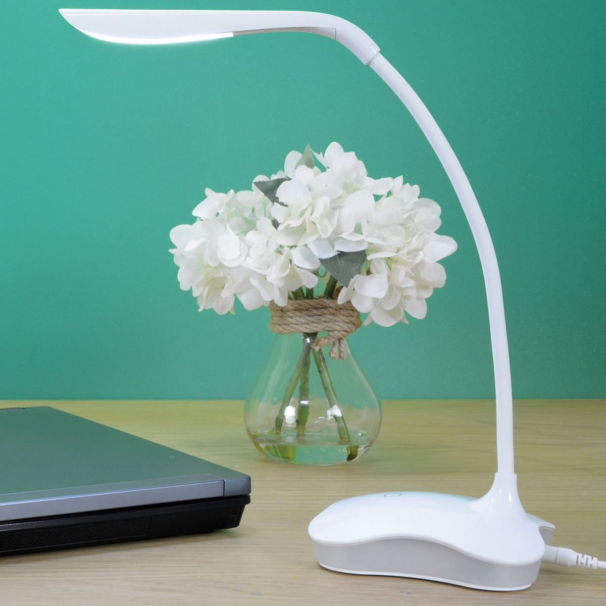 LYYT LED touch sensor USB desk lamp in white with flexible gooseneck illuminated on a wooden desk beside a laptop and a vase of white hydrangeas.