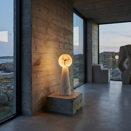 A modern room with large glass windows, concrete walls and floor, features the Talorin 1 Light Sphere Column Alabaster Table Lamp - Soft White on a pedestal. Outside are rocky terrain and sky, while a sculptural artwork stands by another window.