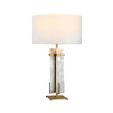 The Pilaster 1 Light Column Alabaster Table Lamp in brass and soft white features a modern alabaster column with gold accents, a large white drum shade, and a gold base. When lit, it emits a soft, warm glow.