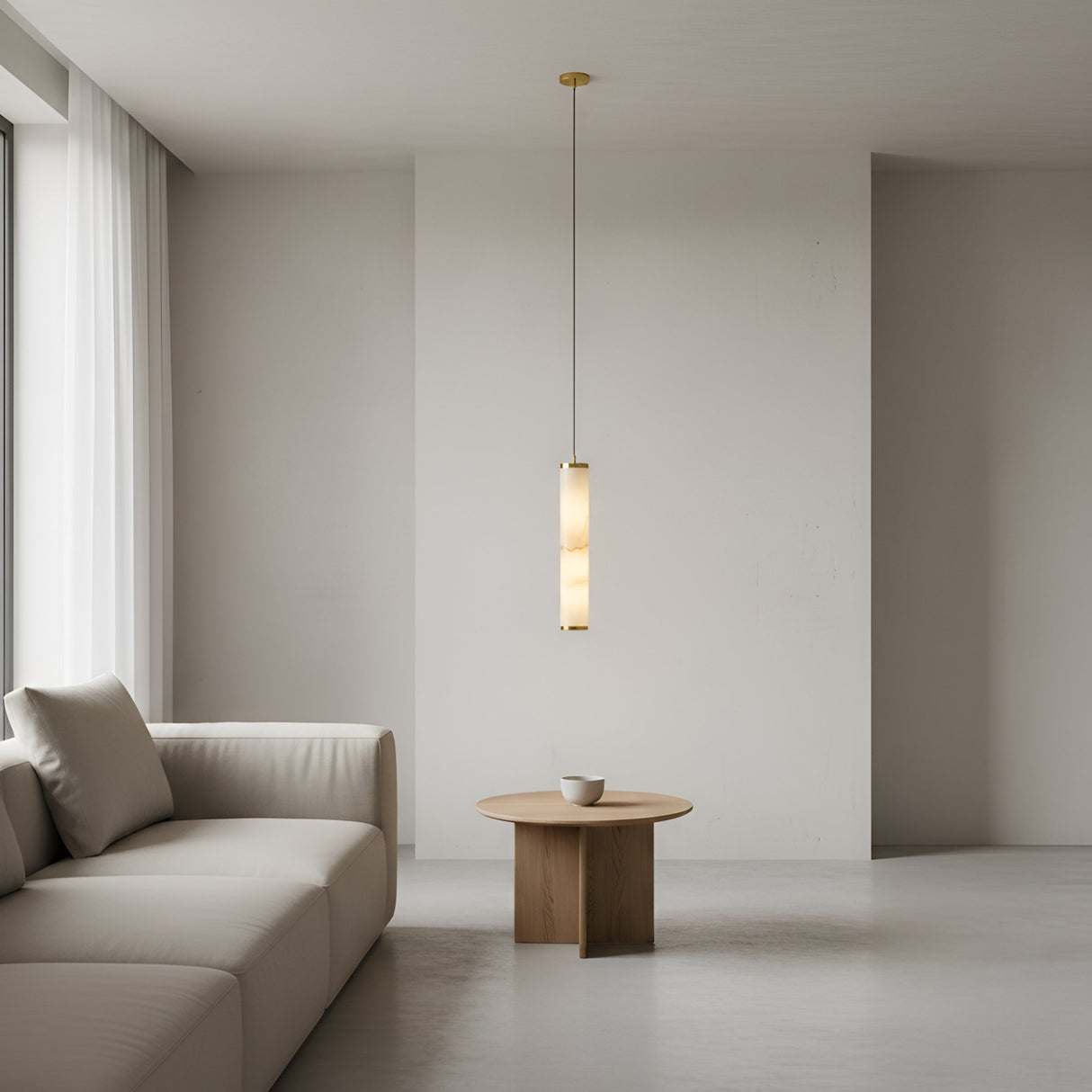 A minimalist living room features a beige sofa, a round wooden coffee table with a white cup, and the Oria LED Medium Alabaster Single Pendant Light in Soft White & Brushed Brass. White walls and sheer curtains on large windows add to the airy atmosphere.