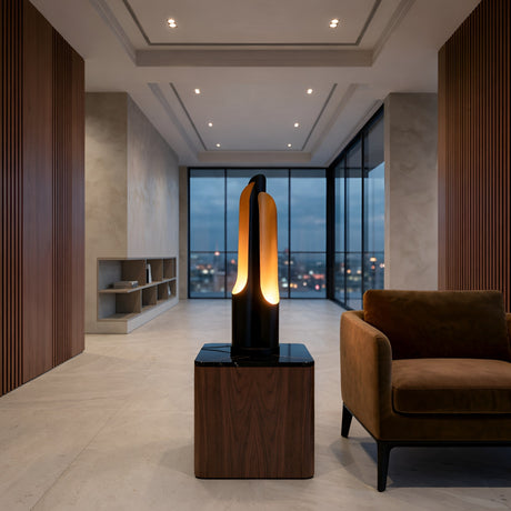 A modern, well-lit living room with large windows, a brown armchair, wood panel accents, and the Stealth Table Lamp - Brass & Black adding warmth beside a sculptural floor lamp with glowing orange light. City lights shimmer outside at dusk.