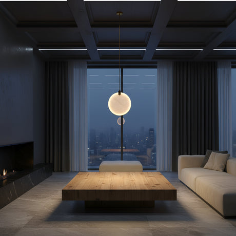 A modern living room at dusk features a large wooden coffee table, cream sofa, minimalist fireplace, and the Luna LED Single Alabaster Pendant Light—Soft White & Brushed Brass—glowing softly in front of a window with city views.