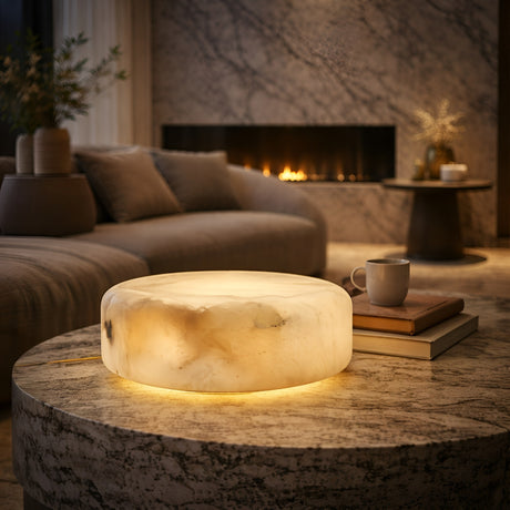 A cozy living room features the Valenor 1 Light Disc Alabaster Table Lamp - Soft White glowing on a marble coffee table with books and a cup nearby. A plush sofa and lit fireplace add warmth to the inviting space.