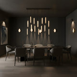 A modern dining room with a dark palette features a long black table, eight grey chairs, abstract wall art, and Marea LED Alabaster Wall Lights in brushed brass above the table, creating a warm, elegant ambiance.