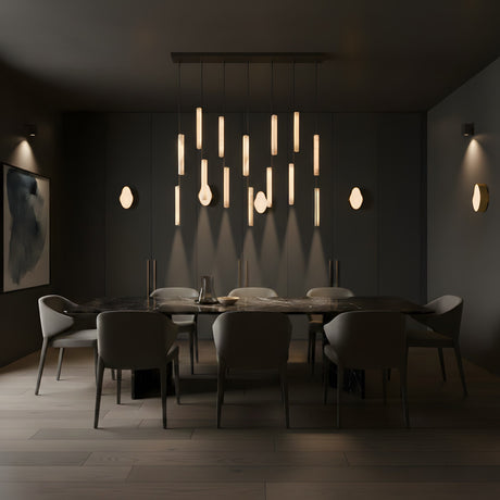 A modern dining room with a dark palette features a long black table, eight grey chairs, abstract wall art, and Marea LED Alabaster Wall Lights in brushed brass above the table, creating a warm, elegant ambiance.