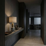 A modern, dimly lit hallway with dark walls and floors features a floating stone shelf adorned with the Lunaro 1 Light Marble Table Lamp in Brass & Black, a decorative vase, and tray. The hallway leads to a naturally lit room.