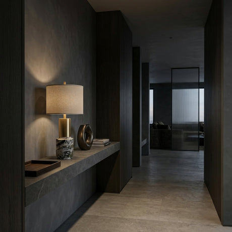A modern, dimly lit hallway with dark walls and floors features a floating stone shelf adorned with the Lunaro 1 Light Marble Table Lamp in Brass & Black, a decorative vase, and tray. The hallway leads to a naturally lit room.