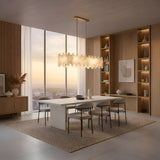 Modern dining room with a long white table, eight gray chairs, the Zarelli 6 Light Interlocking Panel Alabaster Linear Chandelier in Soft White & Brass, built-in wooden shelves, large city-view windows, and neutral tones.