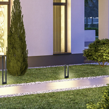 Reno 100cm LED Outdoor Lamp Post in anthracite, a slim open-frame aluminium post light illuminating a gravel-edged garden path beside a modern house at dusk.