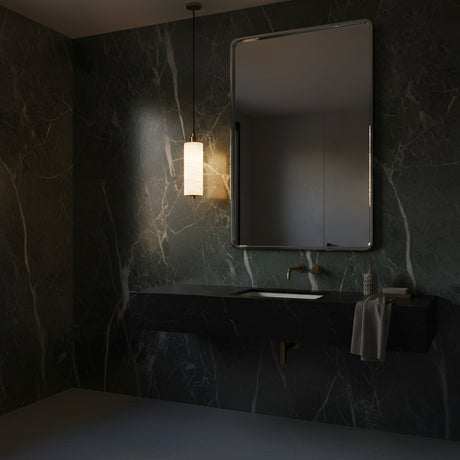 A modern bathroom with dark marble walls, a rectangular mirror, black sink, gold faucet, towel, and the Ardent LED Small Column Alabaster Single Pendant Light in Soft White & Brushed Brass casting a warm glow.