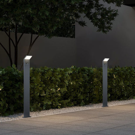 Pearl 100cm LED PIR lamp post in anthracite, slim square post with angled head, installed beside a hedge on a paved outdoor path at night.