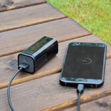AVSL Mercury Emergency USB Power Bank charging a smartphone displaying 57% via black cable on a wooden slatted outdoor table beside grass.