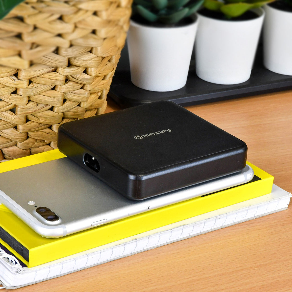 Mercury 65W GaN charger in matt black resting on a smartphone and stacked notebooks beside a woven basket and potted plants on a desk.