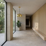 A minimalist hallway with large windows, light stone floors, a built-in wooden bench, a potted plant, and modern decor. The space is bright and modern, featuring the Gulf Glass Pendant Light - Gold as a stylish centerpiece.