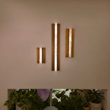 Elvaris LED Large Vertical Alabaster Wall Light in brushed brass, shown lit on a wall in three sizes above potted plants, illuminated stone bar glowing warm white.