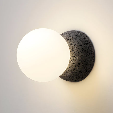 The Origo Concrete Wall Light - Black & White features an opal glass globe diffuser set atop a textured volcanic rock sphere, casting a warm, soft glow against neutral walls.