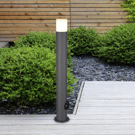Hoosic Tube Floor Lamp in anthracite aluminium, a slim outdoor bollard light with a white diffuser cap, standing on a wooden deck beside garden hedges.