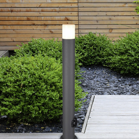 Hoosic 80cm outdoor lamp post in anthracite, slim aluminium column with white diffuser top, set in a garden bed among green shrubs and dark gravel near a wooden deck.