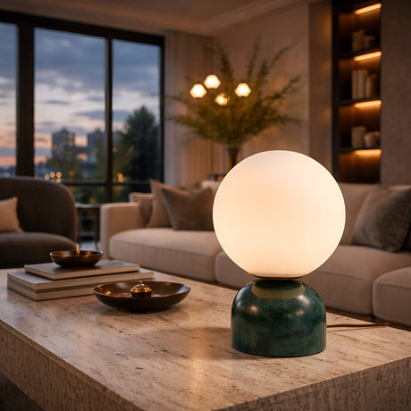 A modern living room with a marble coffee table showcases the Verdane LED Globe Alabaster Table Lamp in soft white and green. A beige sofa, decorative shelves, and large windows revealing a city skyline at dusk complete the elegant space.