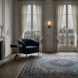 Elegant living room with tall windows, sheer curtains, ornate rug, and a dark blue armchair beside a round table. The Lumara 1 Light Tube Alabaster Wall Light in soft white & brushed brass casts a cozy glow while Parisian rooftops are visible outside.