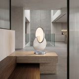 The Half Moon LED Table Lamp - Silver sits on a stone table in a minimalist interior with neutral tones, wood and stone surfaces, glass railing, and concrete walls.