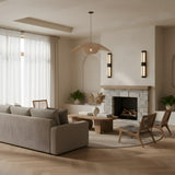A modern living room with neutral tones features a beige sofa, wooden chairs, a coffee table, a stone fireplace, potted plants, sheer-curtained windows, and Caldera LED Small Split-Core Alabaster Wall Lights in matte black & soft white.