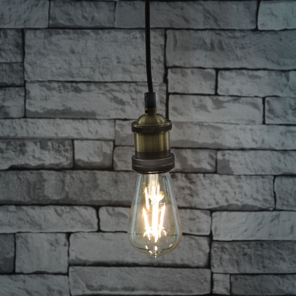 LYYT ES Pendant Set by AVSL in antique brass with knurled E27 lamp holder, braided cord and lit filament bulb hanging against a white brick wall.