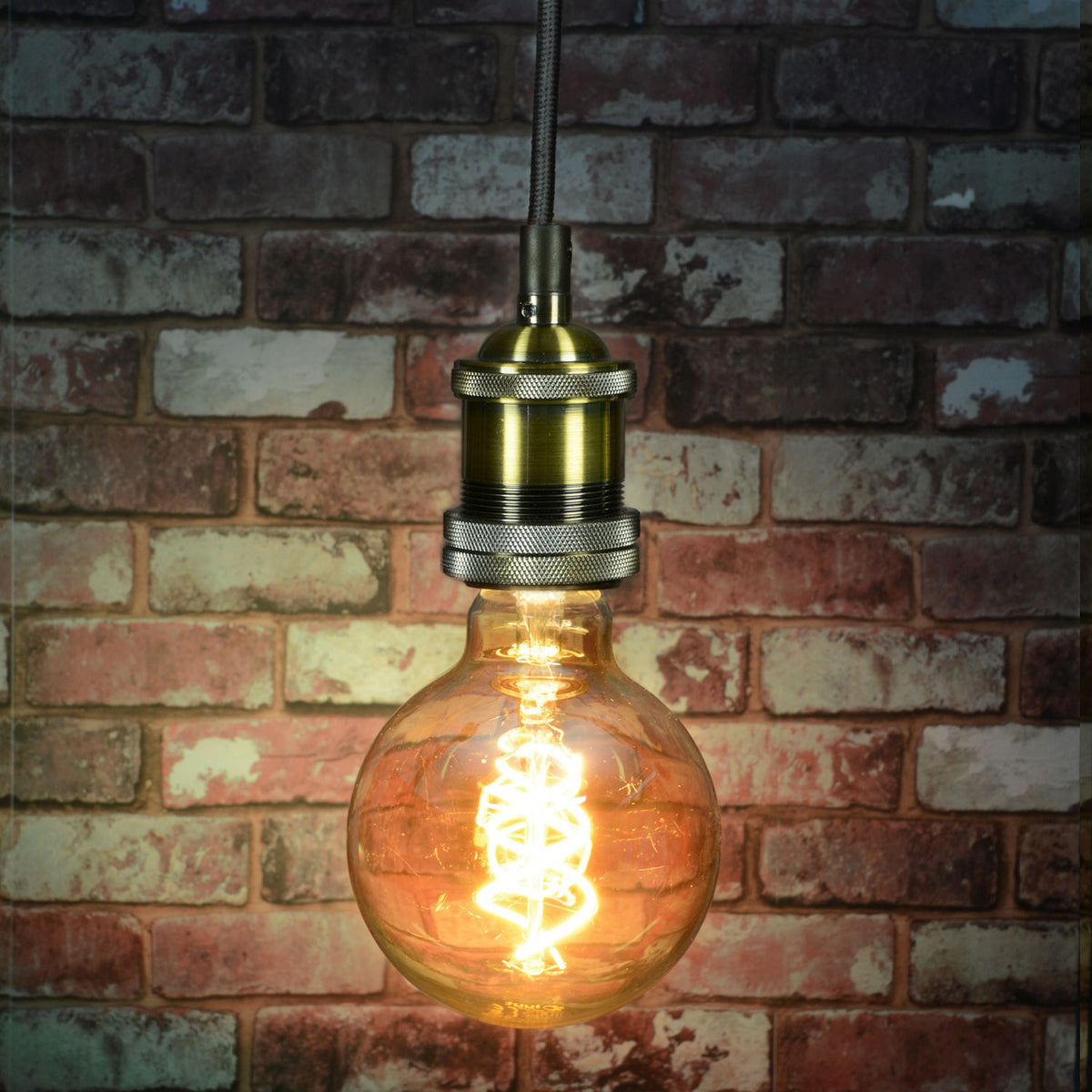 LYYT ES pendant set in antique gold with knurled lamp holder and braided cable, lit globe filament bulb, against an exposed brick wall.