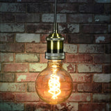 LYYT ES pendant set in antique gold with knurled lamp holder and braided cable, lit globe filament bulb, against an exposed brick wall.