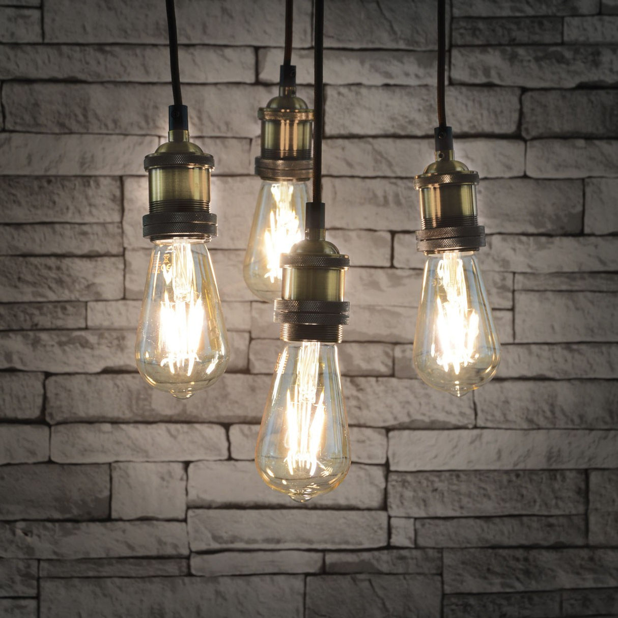 LYYT ES Quad Pendant Set in antique brass with four E27 lamp holders on braided cables, fitted with Edison-style filament bulbs against a grey brick wall.