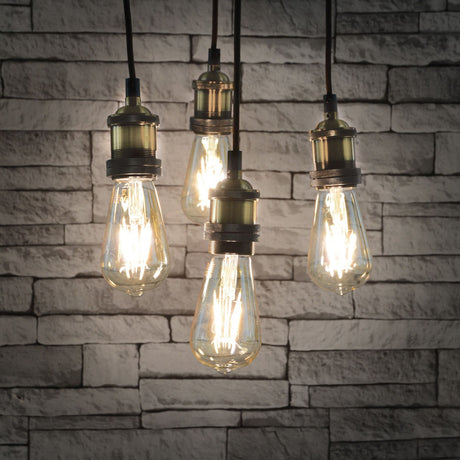 LYYT ES Quad Pendant Set in antique brass with four E27 lamp holders on braided cables, fitted with Edison-style filament bulbs against a grey brick wall.