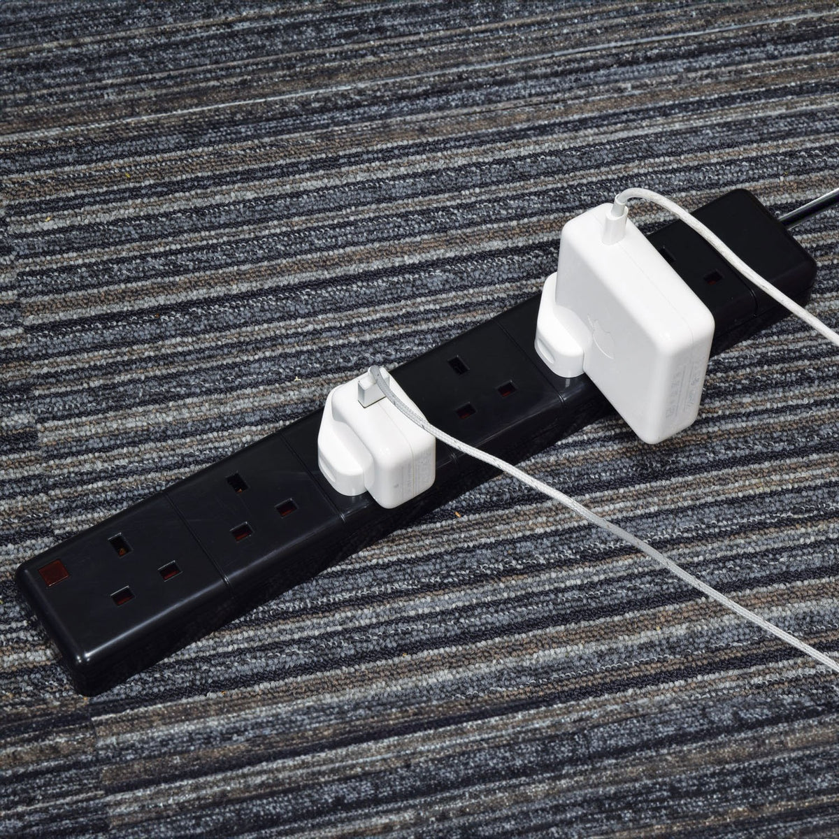 AVSL Mercury 6 Gang 13A trailing socket in black with two white Apple plug adapters connected, shown on a striped grey carpet floor.
