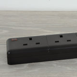 Close-up of the AVSL Mercury 6 Gang 13A trailing socket in black thermoplastic, showing UK 3-pin outlets and a red neon indicator on a light wood surface.