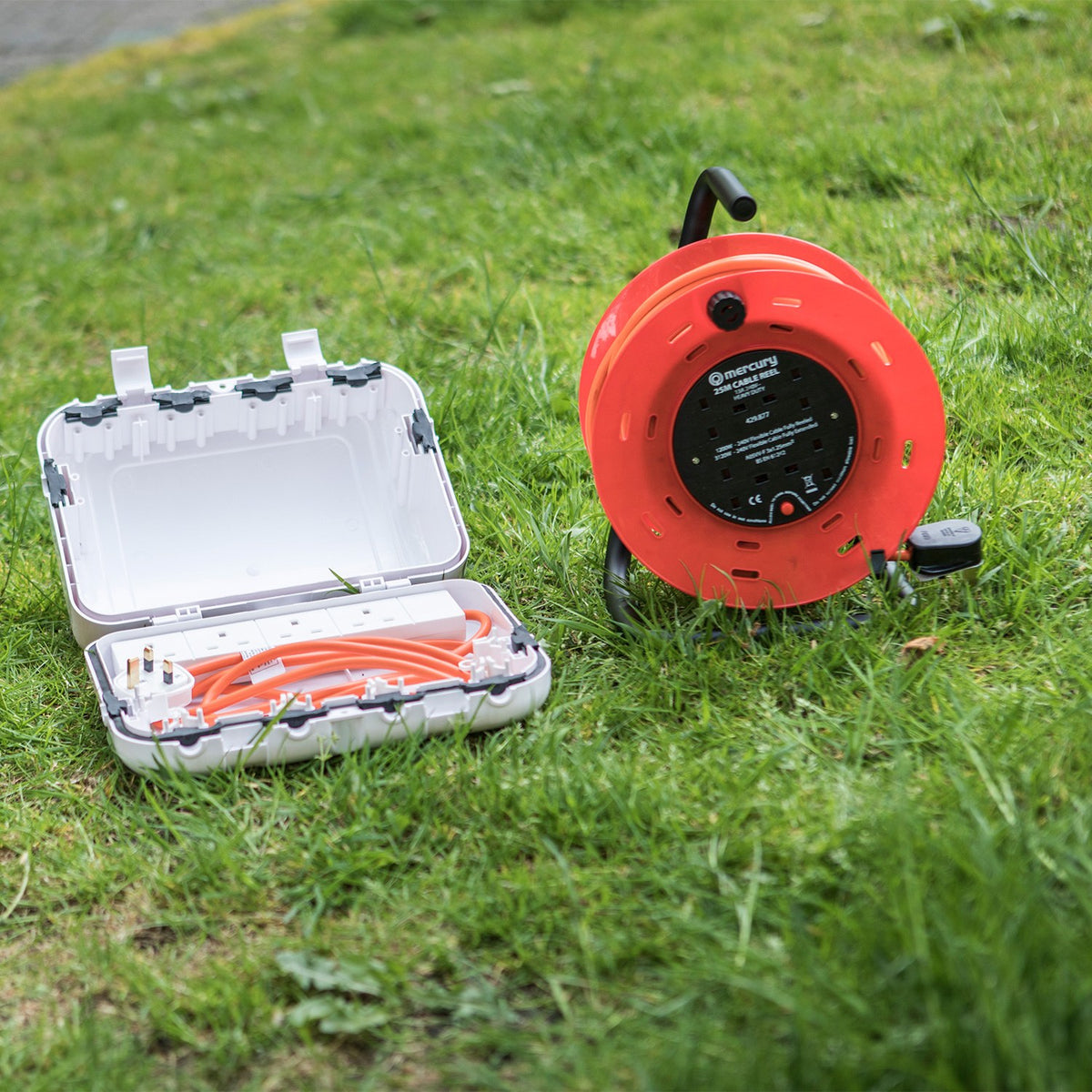 Mercury 25m 4 gang cable reel in orange with steel frame and carry handle, shown on grass beside an open weatherproof extension lead box.