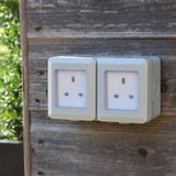 AVSL Mercury IP55 2 gang weatherproof socket in white with transparent hinged covers over twin UK outlets, surface-mounted on a weathered timber wall.