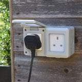 AVSL Mercury IP55 2 gang weatherproof white socket mounted on a timber wall, one hinged cover open with a UK plug inserted.
